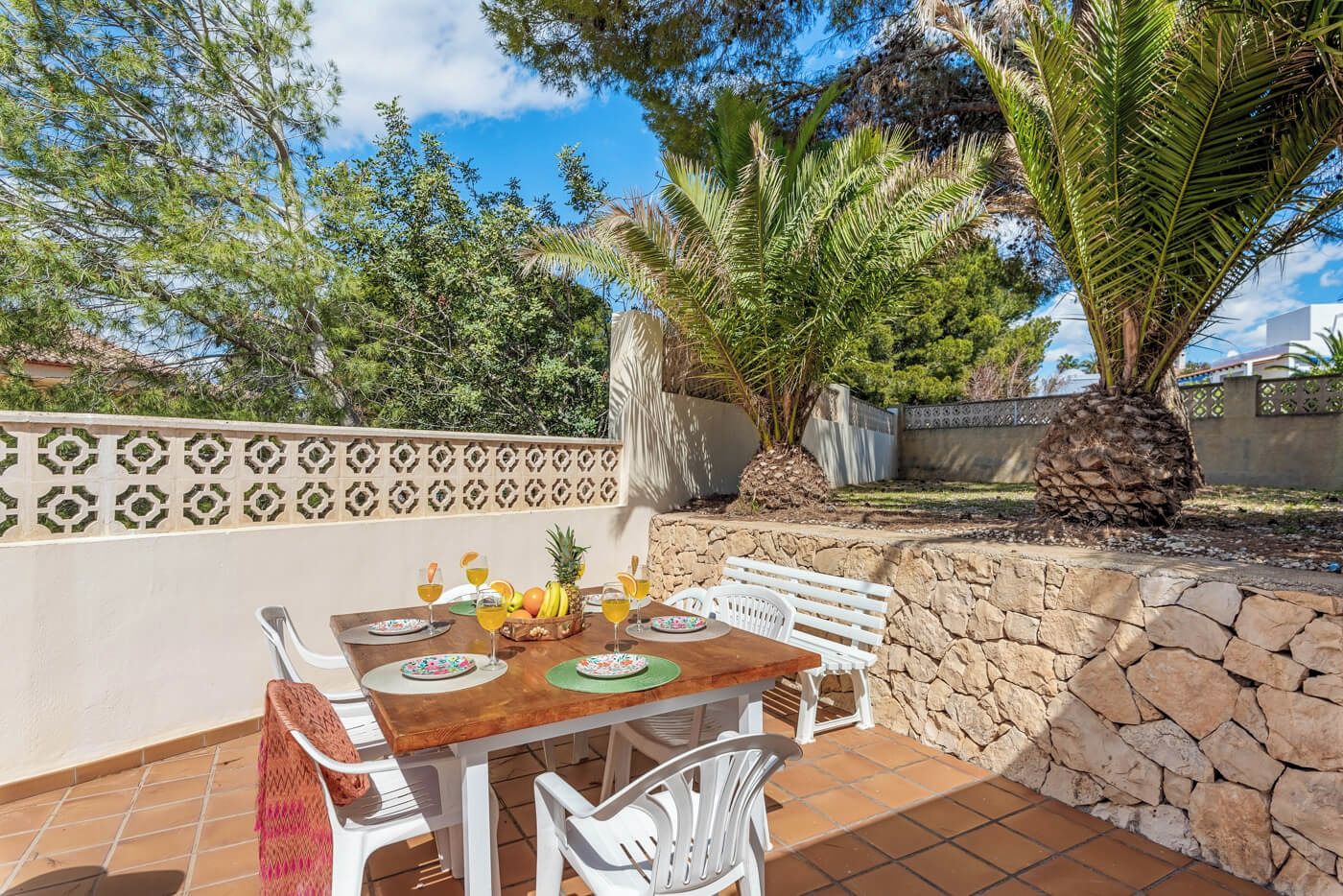 En udendørs terrasse med et træbord dækket til spisning, omgivet af hvide stole og en stenvæg.