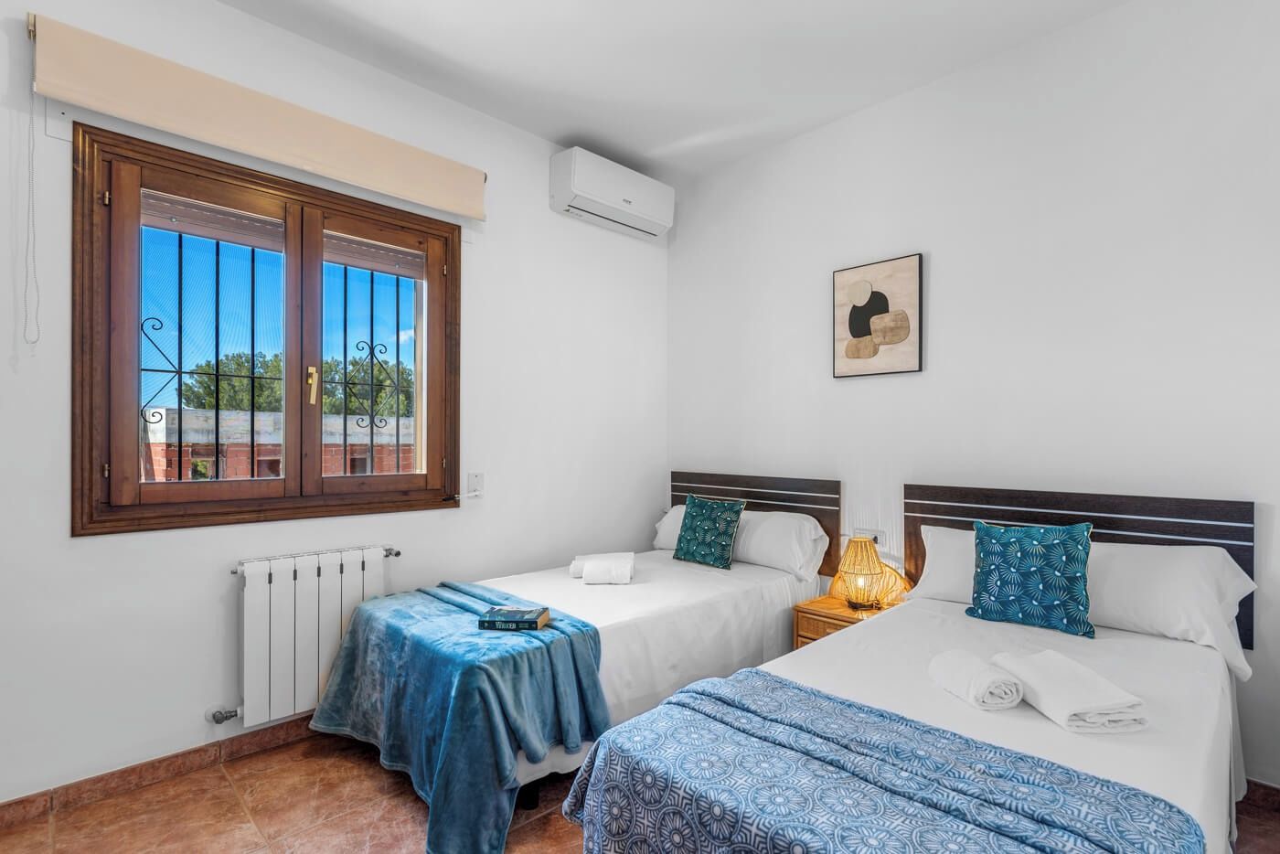 Quarto de hotel com duas camas, um radiador, uma janela com grades e um ar condicionado fixado na parede.