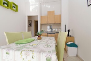 A compact kitchen with wooden cabinets, a dining table with a green table runner, and a trash bin. - Apartments Milenko (Vodice)