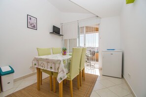 A small dining area with a table set for two, a refrigerator, and a window with curtains. - Apartments Milenko (Vodice)