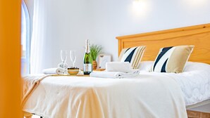 Una cama bien hecha con cabecera de madera, una botella de champán, dos copas y un frutero al lado.