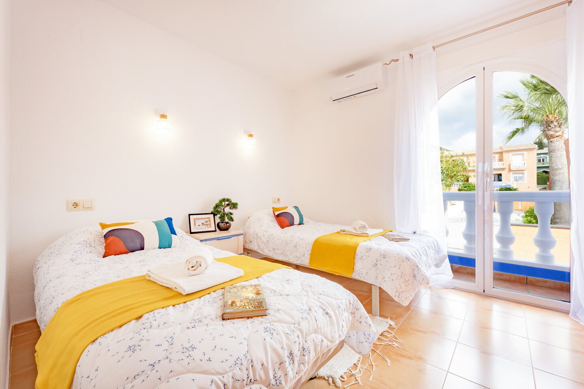 A room with two beds, each with a yellow blanket and a book. There is a window with a view of buildings and palm trees.