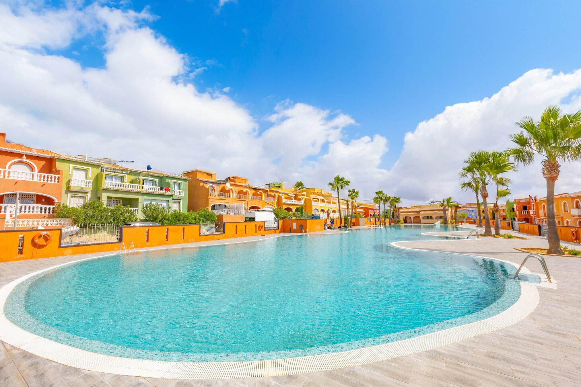 A large, clear swimming pool with a curved edge, surrounded by buildings with balconies and palm trees.