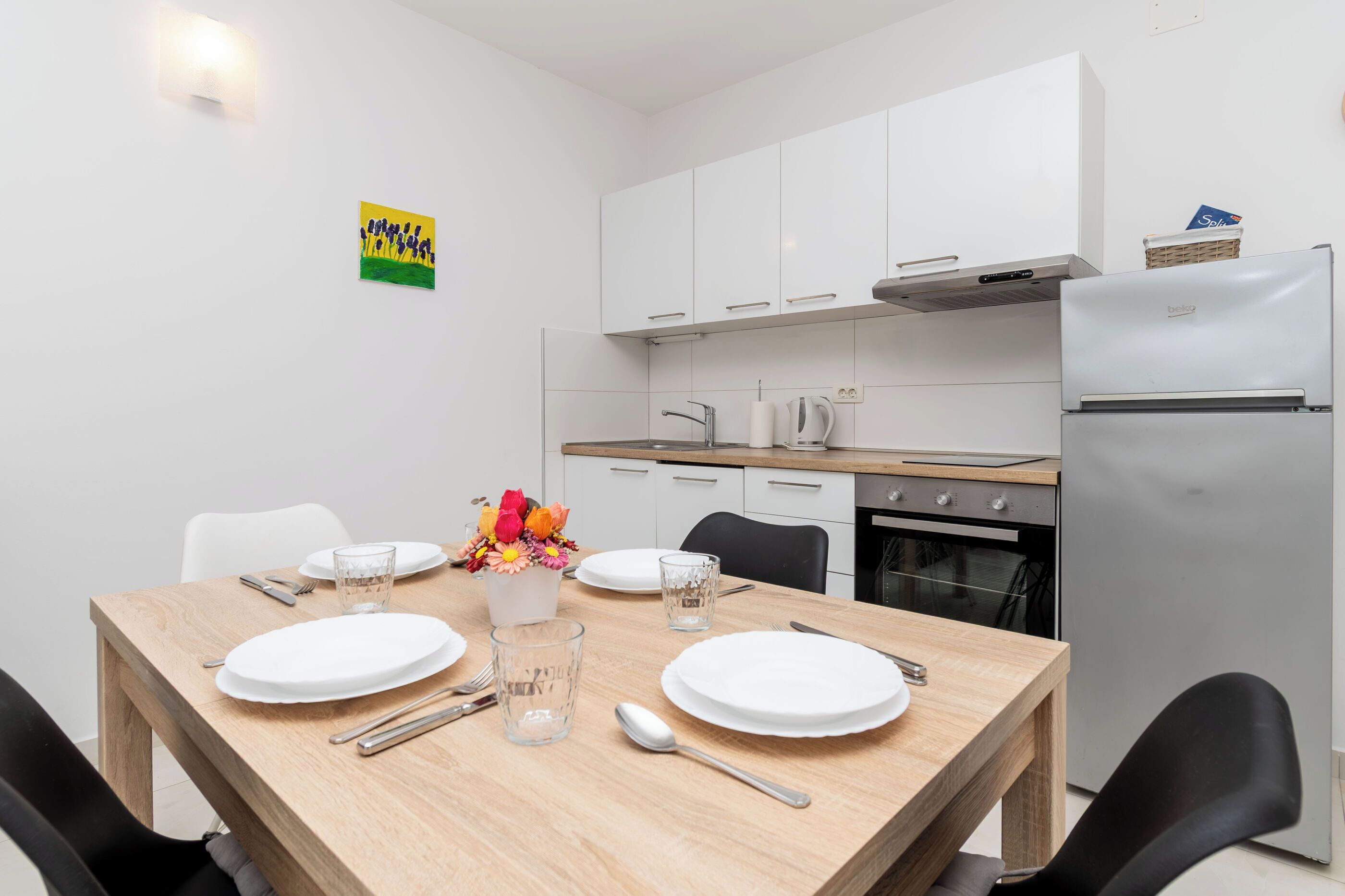 A modern kitchen with white cabinets, a refrigerator, and a dining table set for four with plates, cutlery, and glasses.