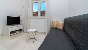 Une chambre avec un lit, un téléviseur posé sur un meuble blanc, une petite table et une fenêtre avec des rideaux.