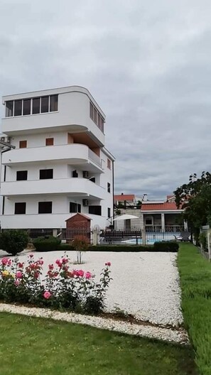 A multi-story white building with balconies, a gated swimming pool, and a well-maintained garden with pink flowers. - House DD With Heated Pool (Okrug)