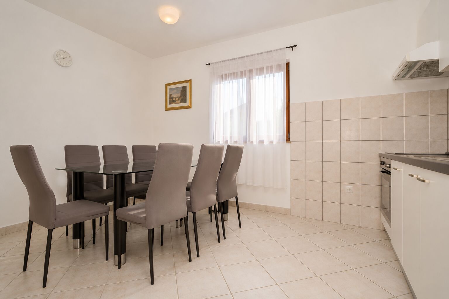 A dining area with a round table and six chairs, a kitchen with white cabinets, and a window with sheer curtains.