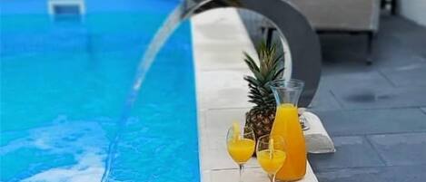 A swimming pool with a water feature, a pineapple, a glass of orange juice, and a bowl of strawberries.