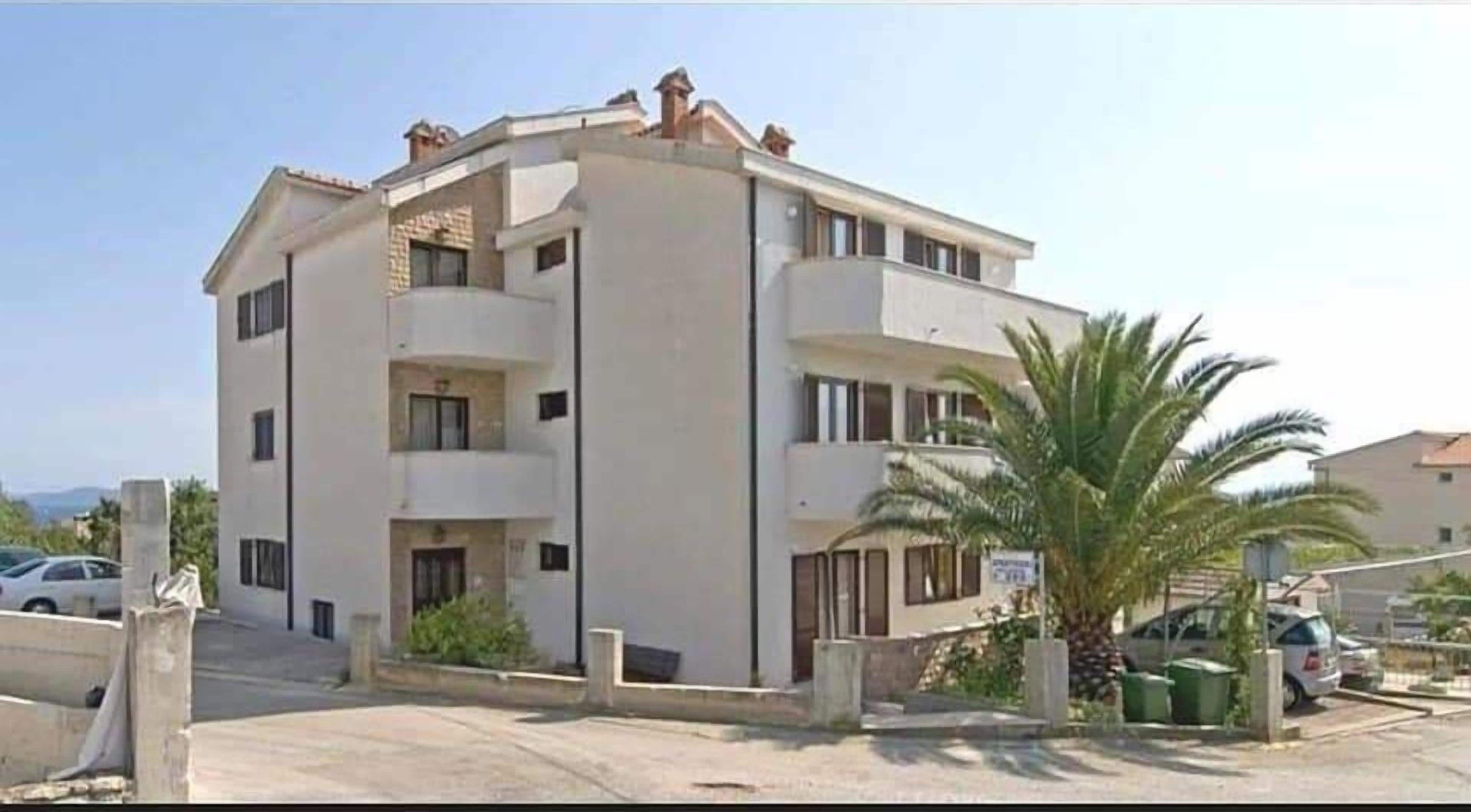A multi-story residential building with a flat roof, balconies, and a parking area with cars.