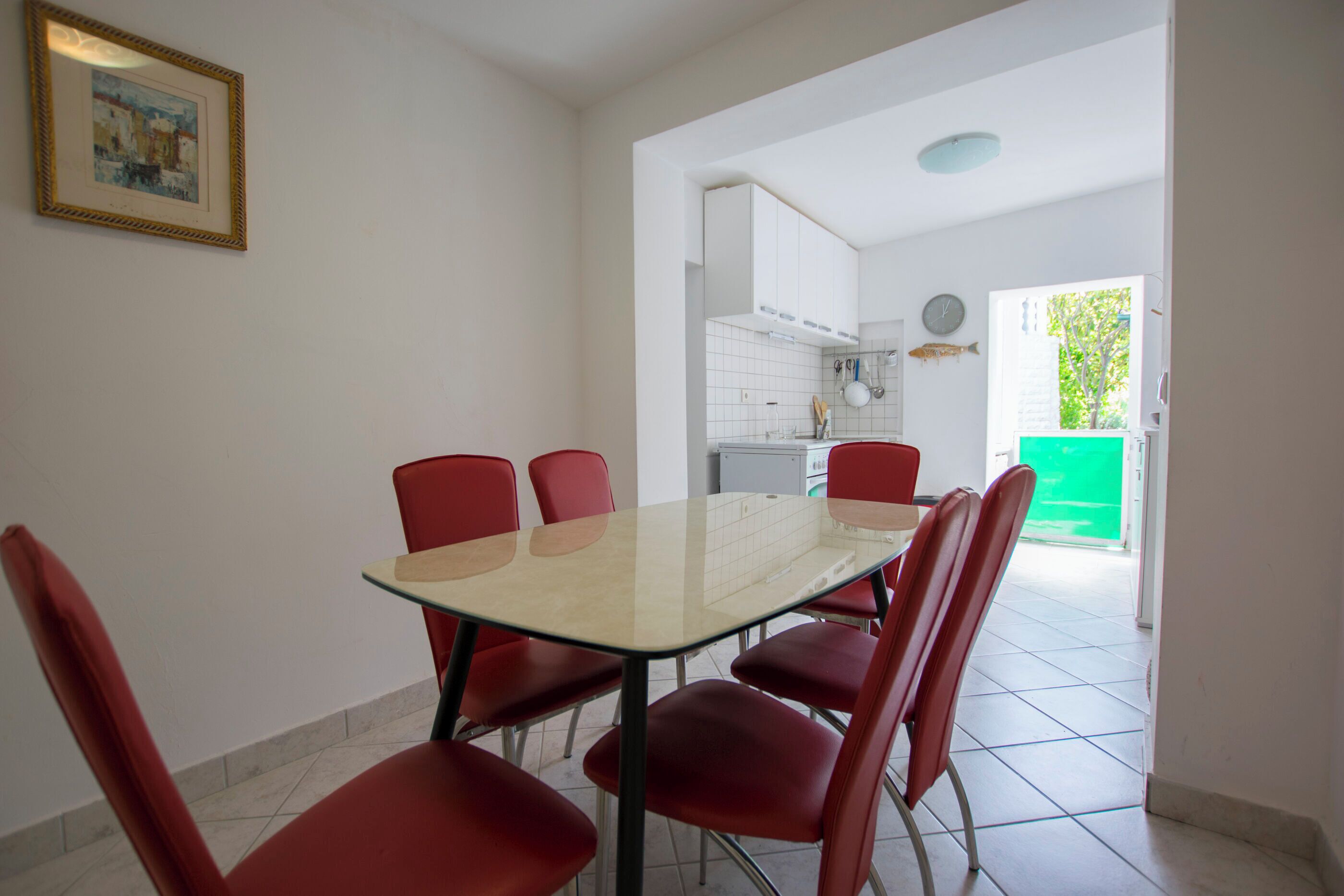A dining area with a glass table and red chairs, a kitchen in the background, and a framed picture on the wall.