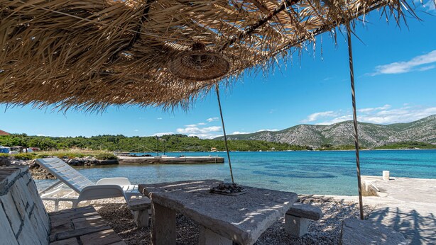 Ein Sitzbereich am Strand mit einem Steintisch und Liegestühlen, mit Blick auf das klare blaue Meer und grüne Hügel.