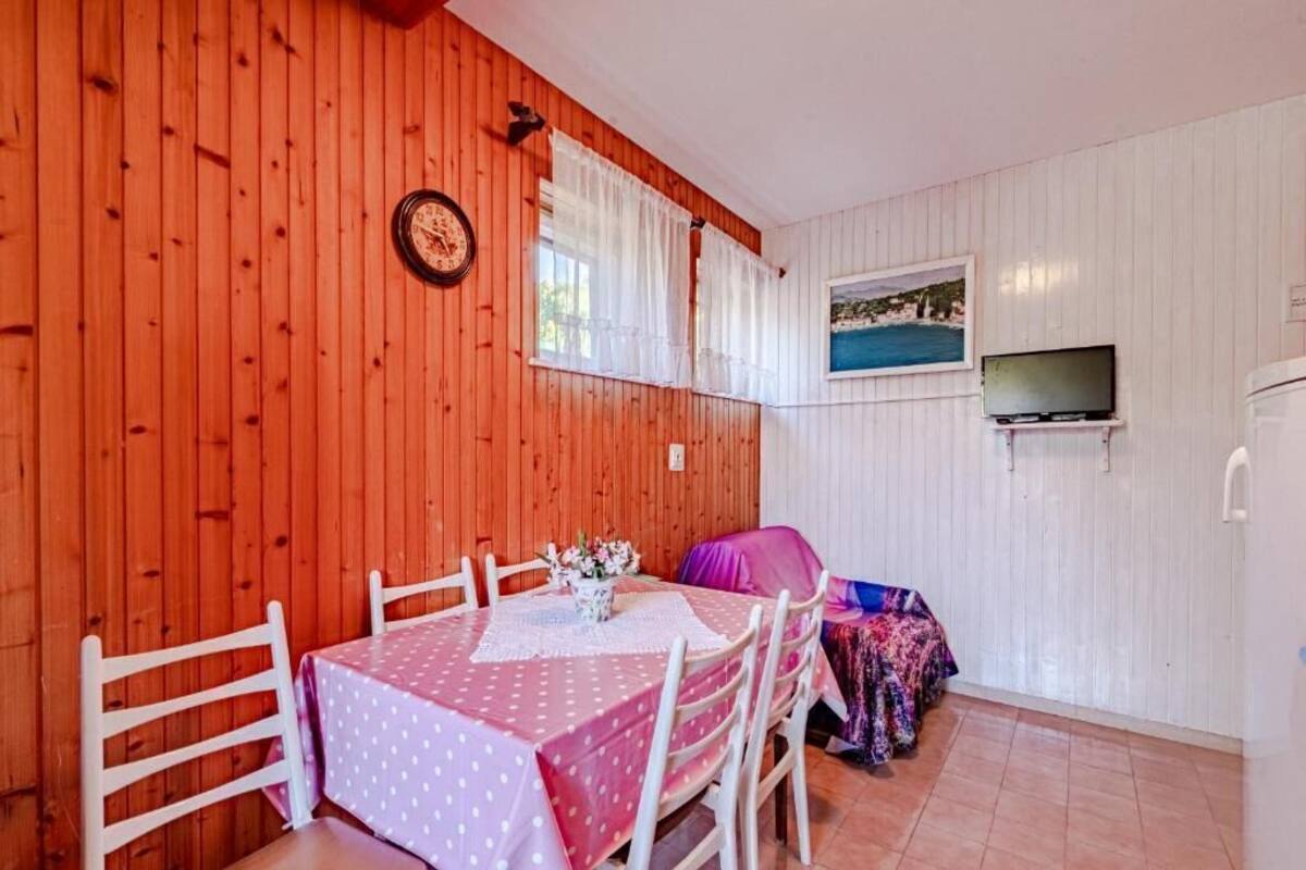 Une pièce chaleureuse aux murs en bois, une table ronde recouverte d’une nappe à pois, des chaises blanches, un lit avec une couverture colorée, un téléviseur et une horloge au mur.