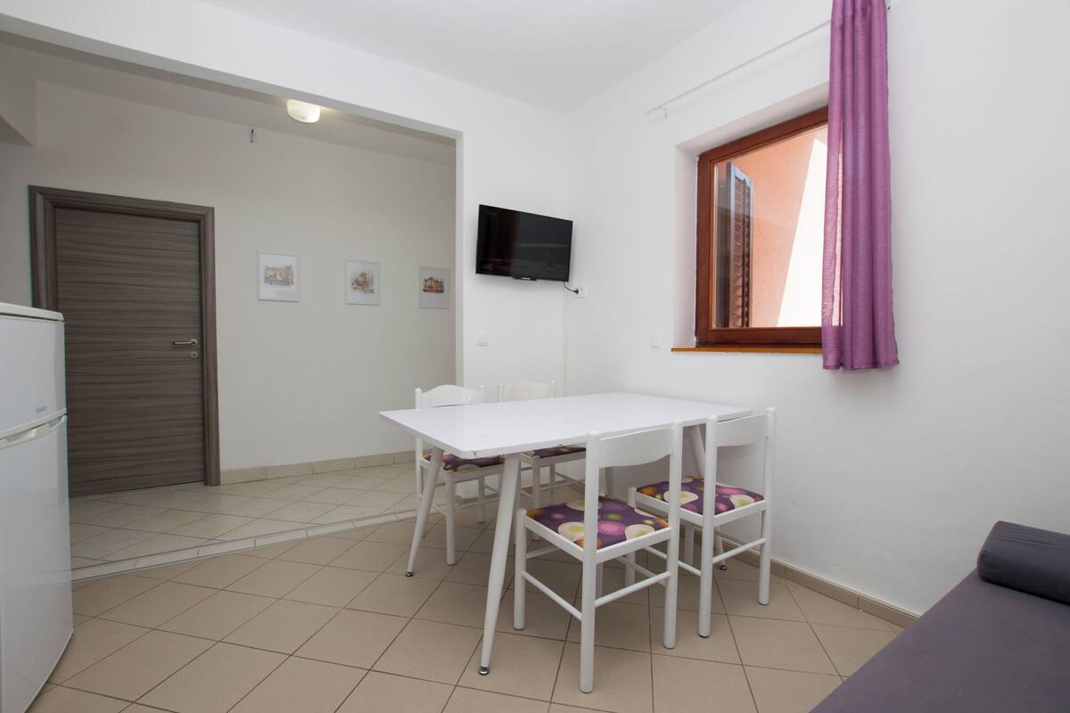 A small, clean room with a white table and chairs, a TV mounted on the wall, and a window with purple curtains.
