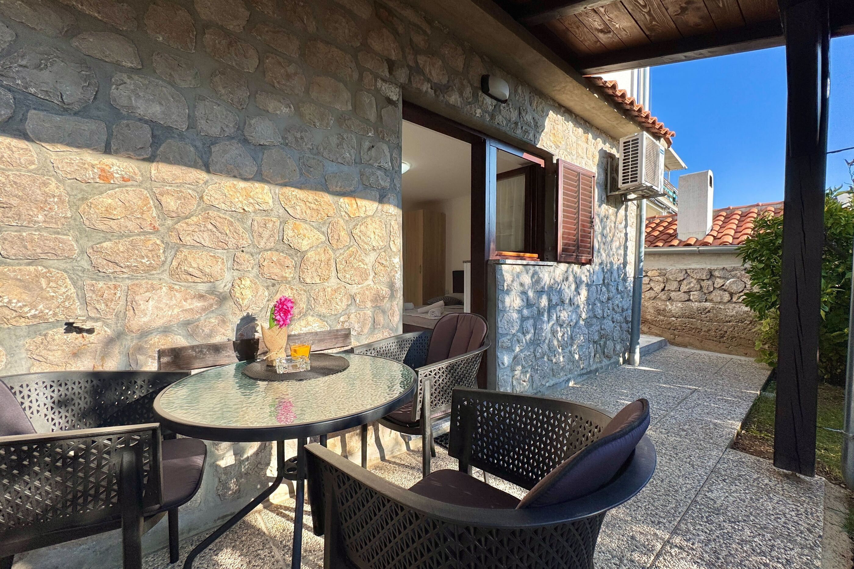 A stone exterior wall, a round glass table with chairs, and a window with shutters.