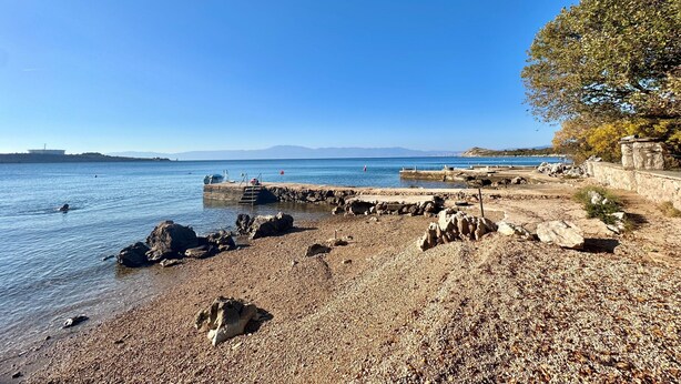 A pebbled beach with a rocky shoreline, a clear blue sky, and distant mountains. - Apartments Matosic (Omisalj)