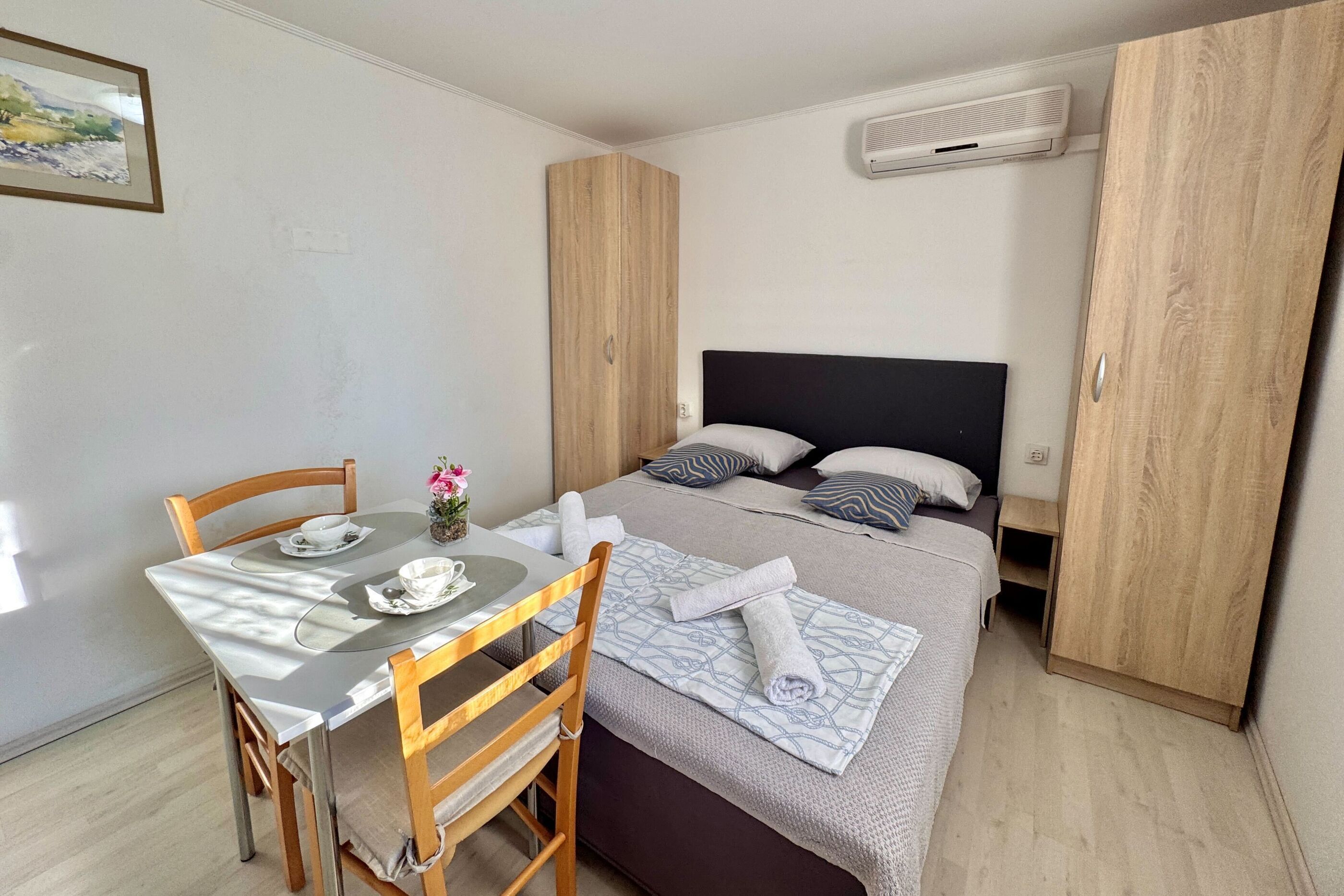 Une pièce bien rangée, avec un lit, une table entourée de chaises et une petite table sur laquelle est posé un vase de fleurs.