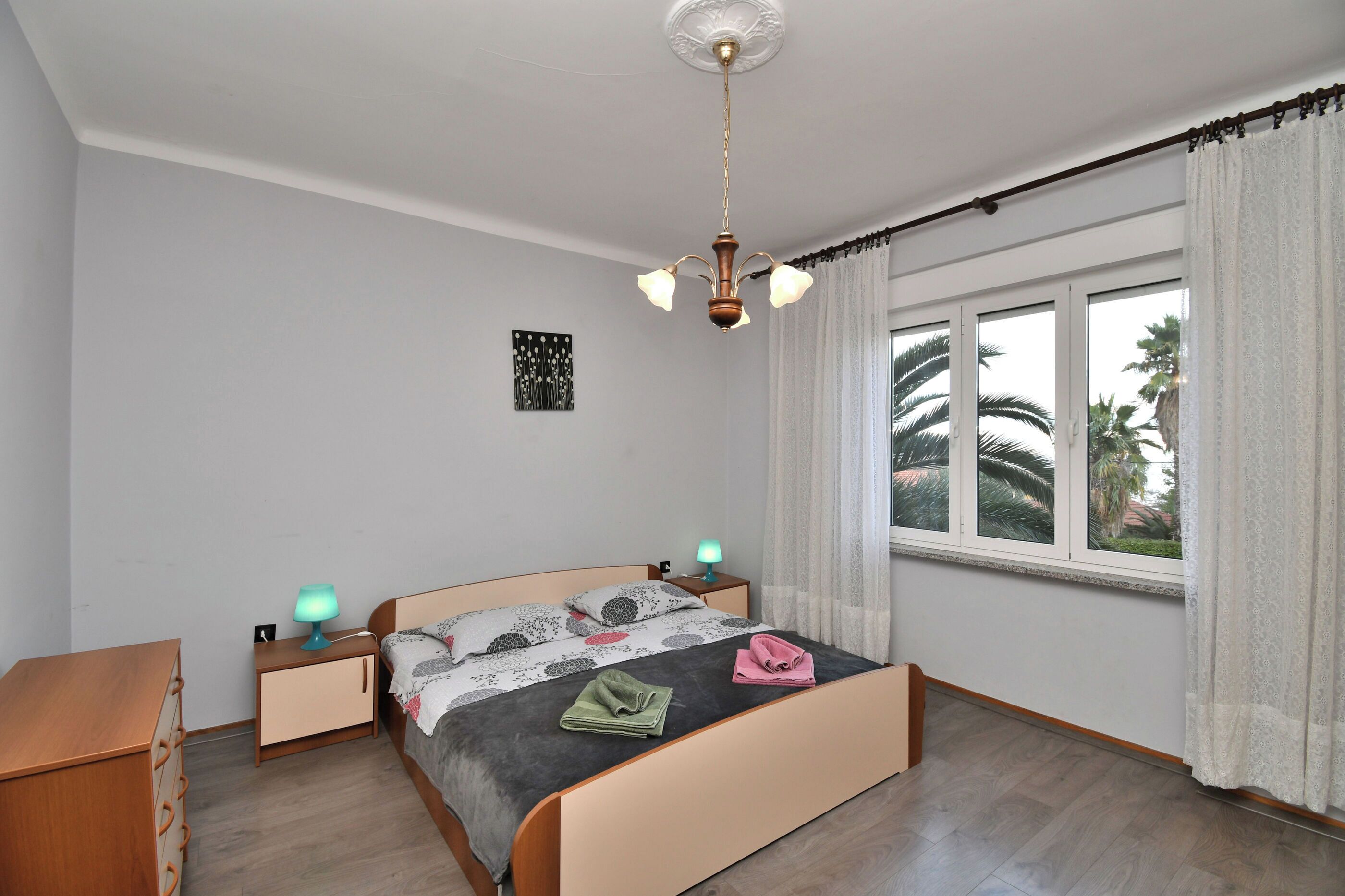 Quarto com cama, mesas de cabeceira, abajur, iluminação no teto, janela com cortinas e uma obra de arte na parede.
