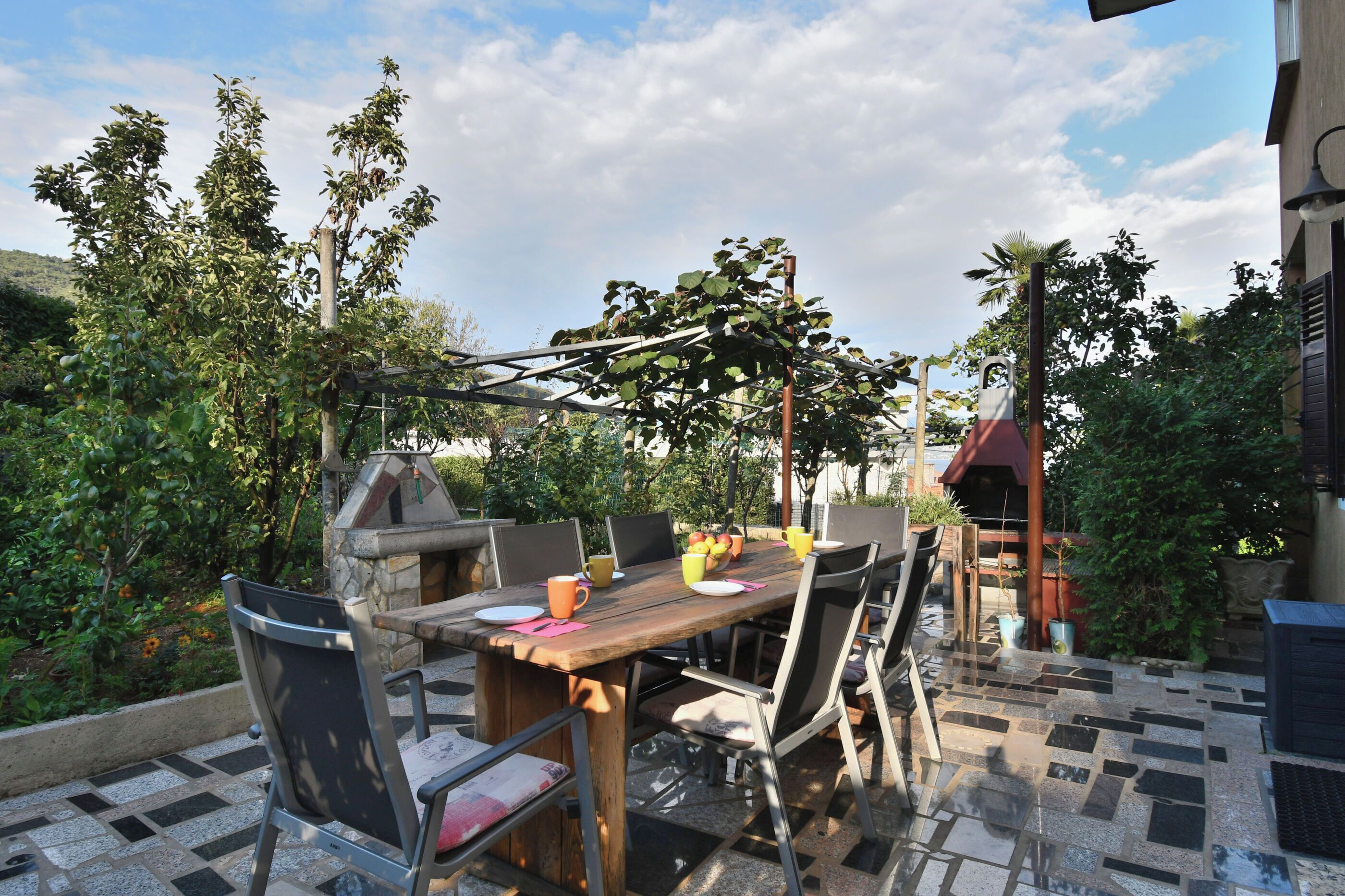 An outdoor dining area with a wooden table and metal chairs, surrounded by greenery and a pergola.