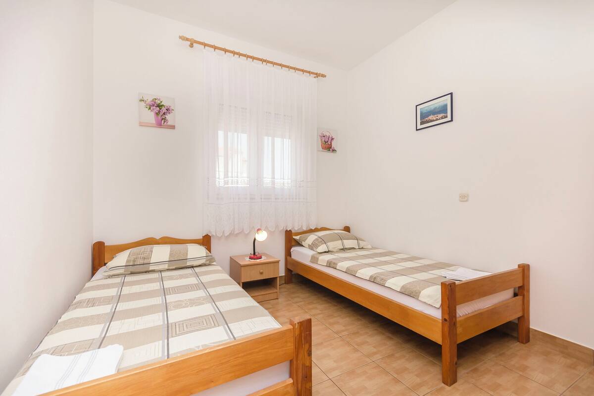 Ein Schlafzimmer mit zwei hölzernen Betten, einem Nachttisch mit Lampe, einem Fenster mit durchsichtigen Vorhängen und einem gerahmten Bild an der Wand.