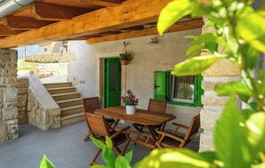 Una scala in pietra conduce a un terrazza in legno con tavolo e sedie, persiane verdi e una pianta pensile.