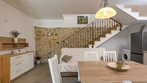 A cozy living area with a sofa, dining table, and a stone wall. A staircase with a metal railing leads to an upper level. - House Casa Della Nonna (Krsan)