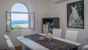 Un coin repas comprenant une table, des chaises, un vase avec des fleurs, un saladier rempli de fruits et une vue sur la mer par une fenêtre ouverte.