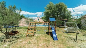 Une aire de jeux avec un toboggan, des balançoires et une structure d’escalade, située dans un espace verdoyant, avec des arbres et des maisons en arrière-plan.