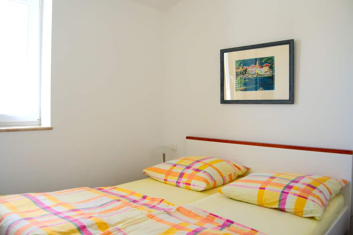 Una cama individual con una colcha a cuadros y dos almohadas, un cuadro en la pared y una ventana a la izquierda.