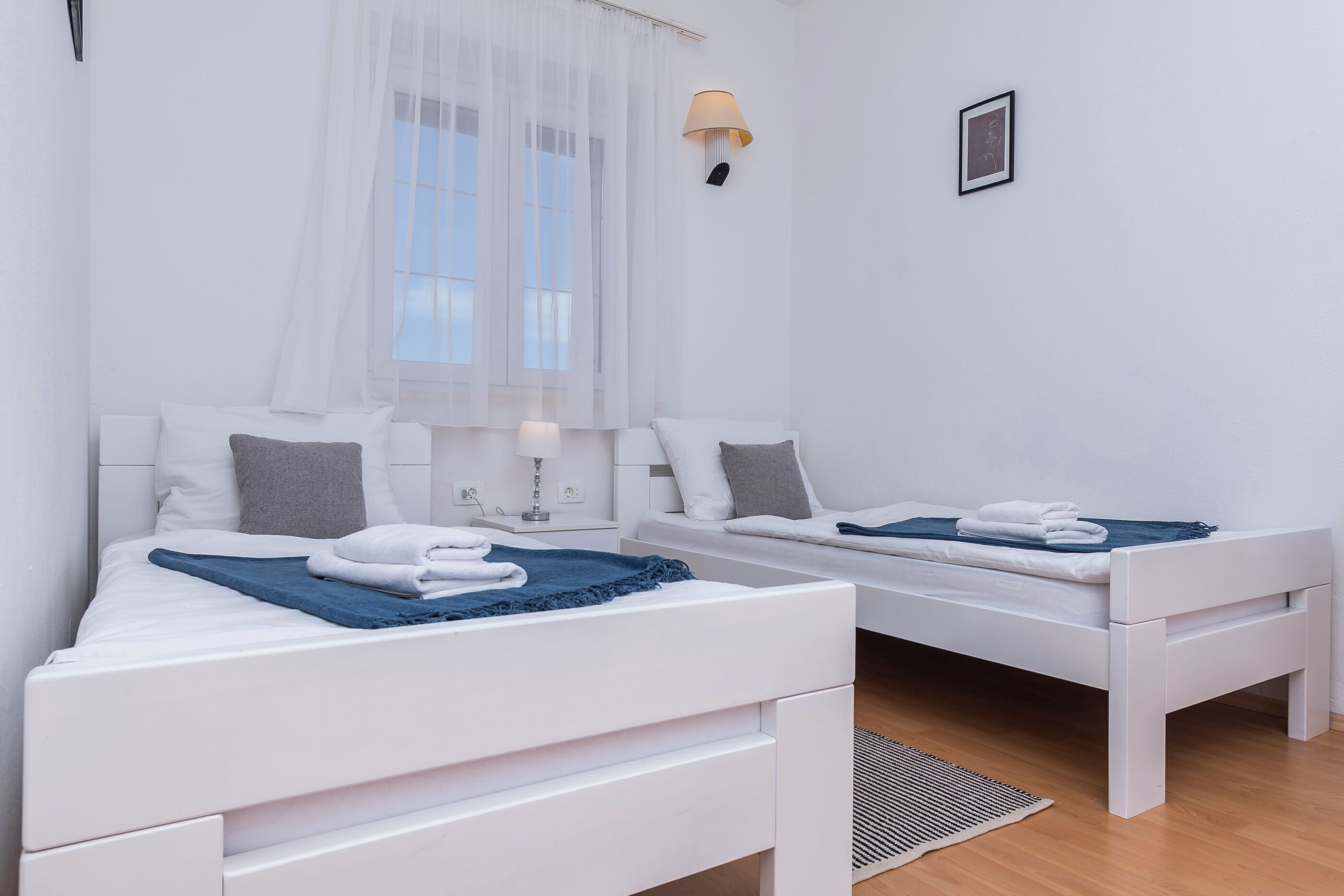 Ett hotellrum med två vita sängar, ett fönster med tunna, genomskinliga gardiner, en vägglampa och en liten tavla i en ram.