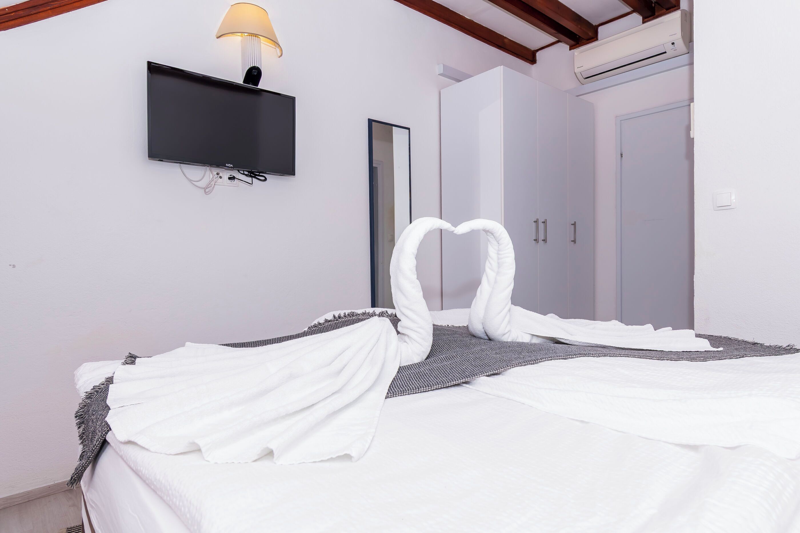 A neatly made bed with white linens and a heart-shaped towel arrangement. A flat-screen TV is mounted on the wall above the bed.