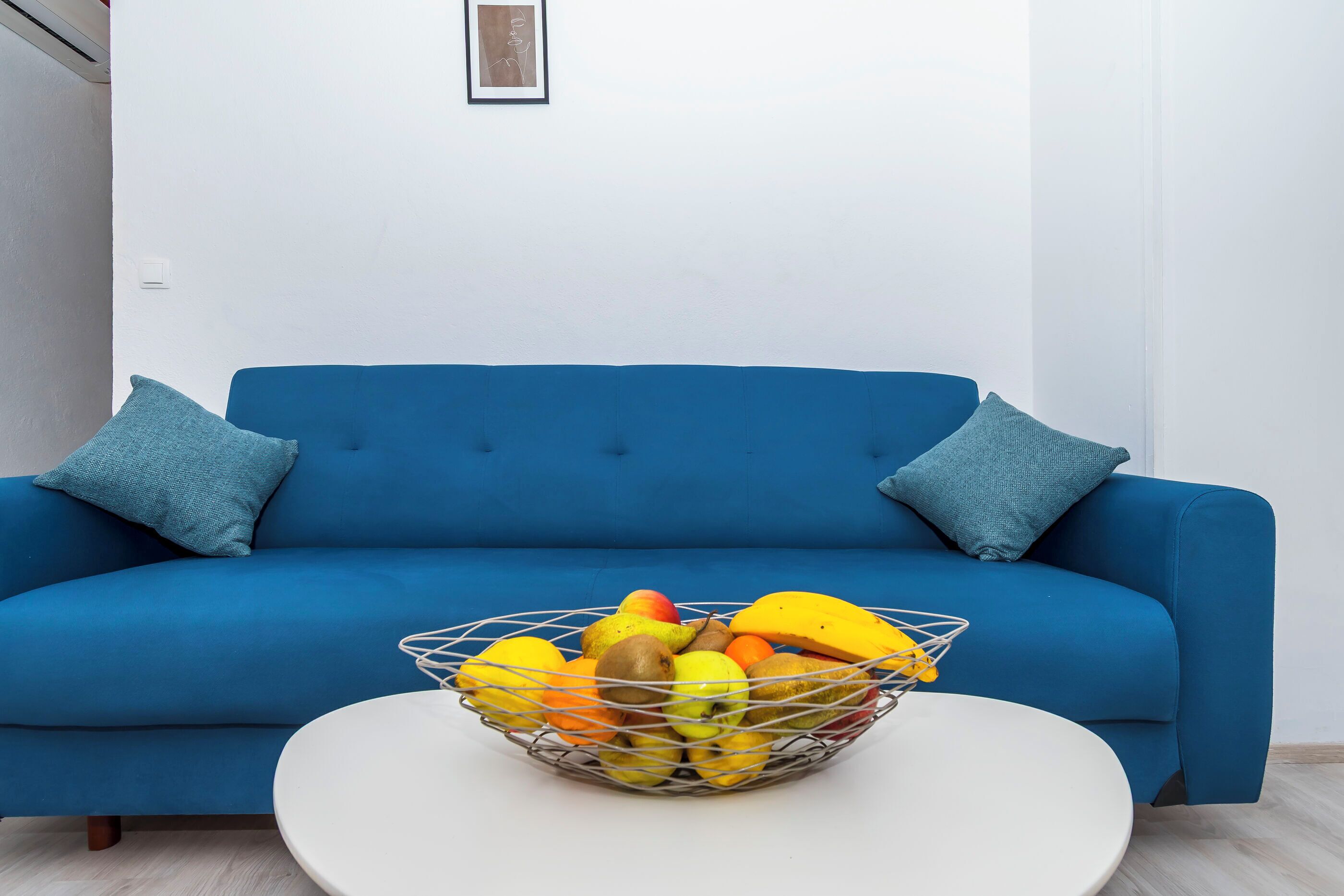 Un sofá azul con dos almohadas, una mesa blanca y una cesta de frutas.