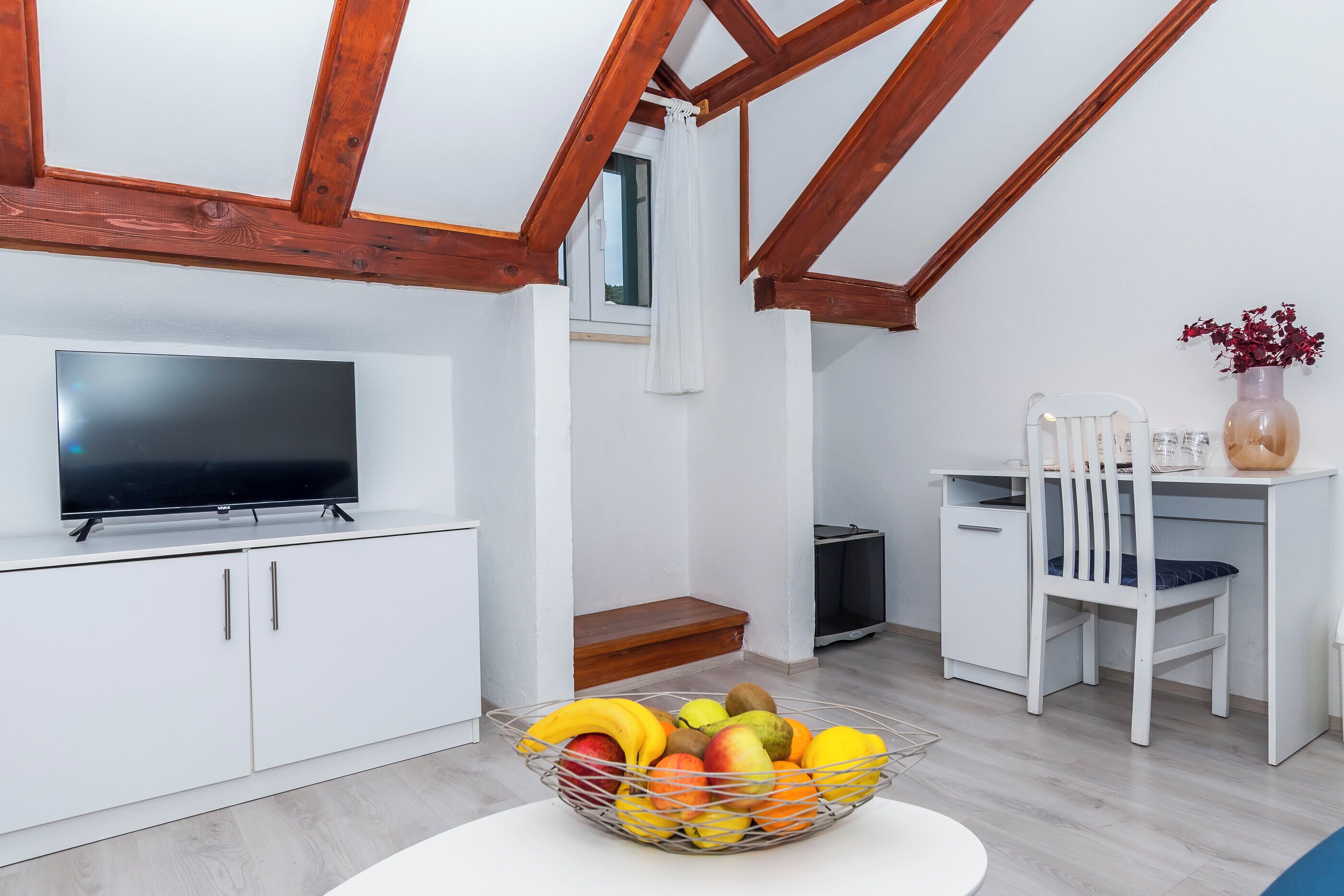 A modern kitchen with a TV, white cabinets, a fruit bowl, and a dining area with a chair and table.