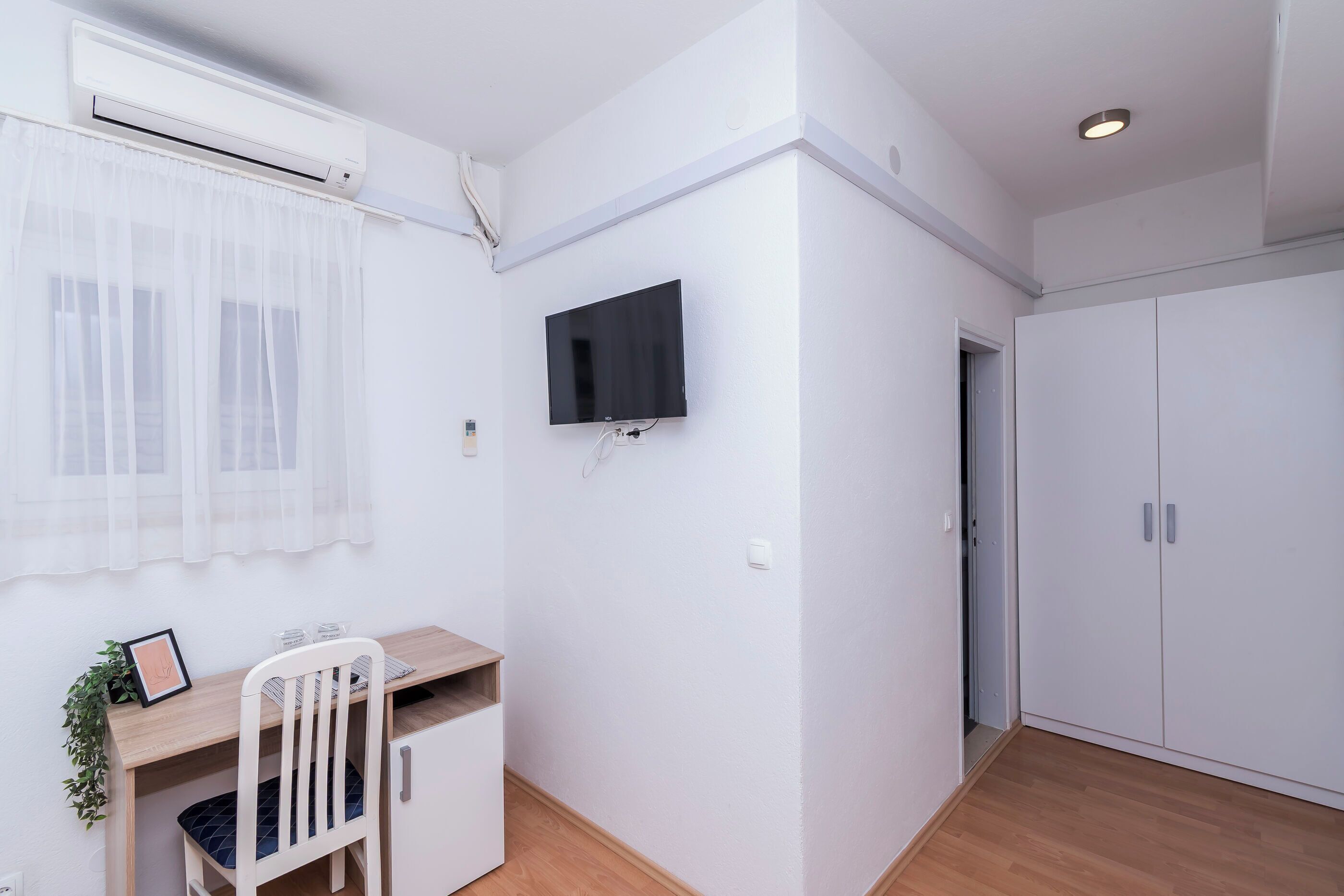 Una habitación con escritorio de madera, una silla blanca, televisor montado y armario.