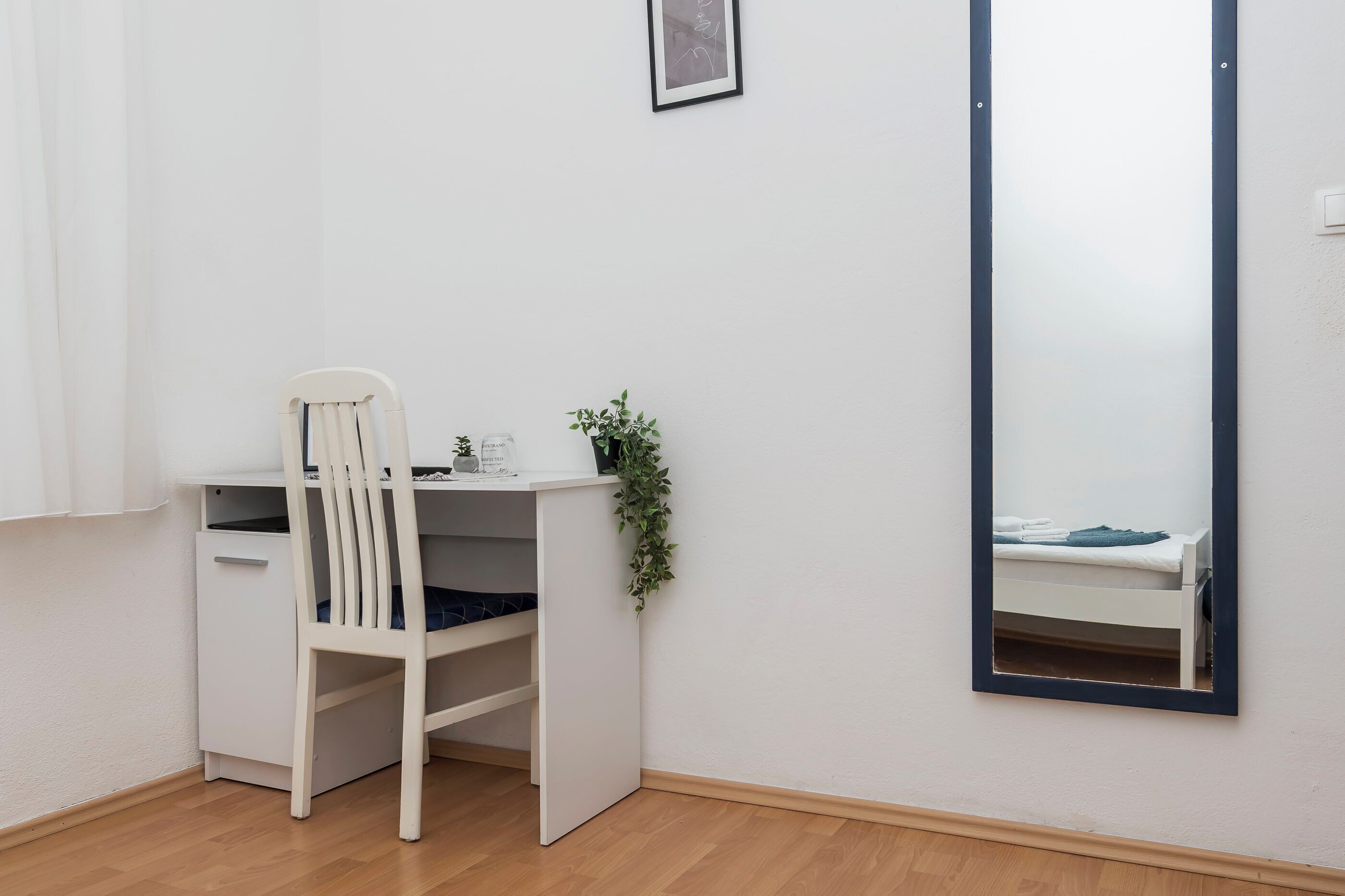 Uma mesa branca com uma cadeira, um pequeno vaso de plantas e um espelho refletindo uma cama.