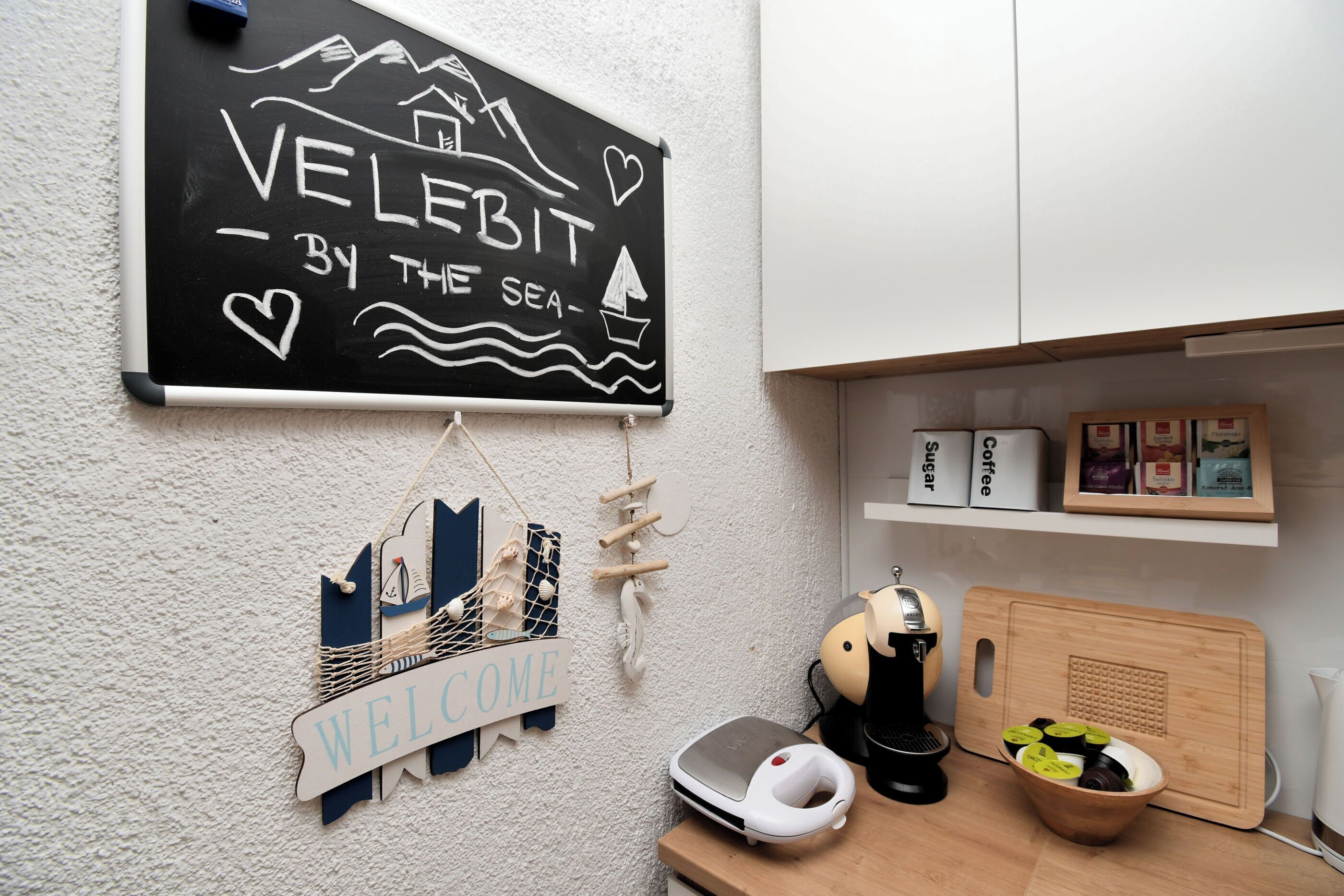 Une cuisine avec un tableau noir sur lequel on peut lire « VELEBIT, AU BORD DE LA MER », un panneau d’accueil, une machine à café, une balance et une planche à découper avec des tranches de kiwi.