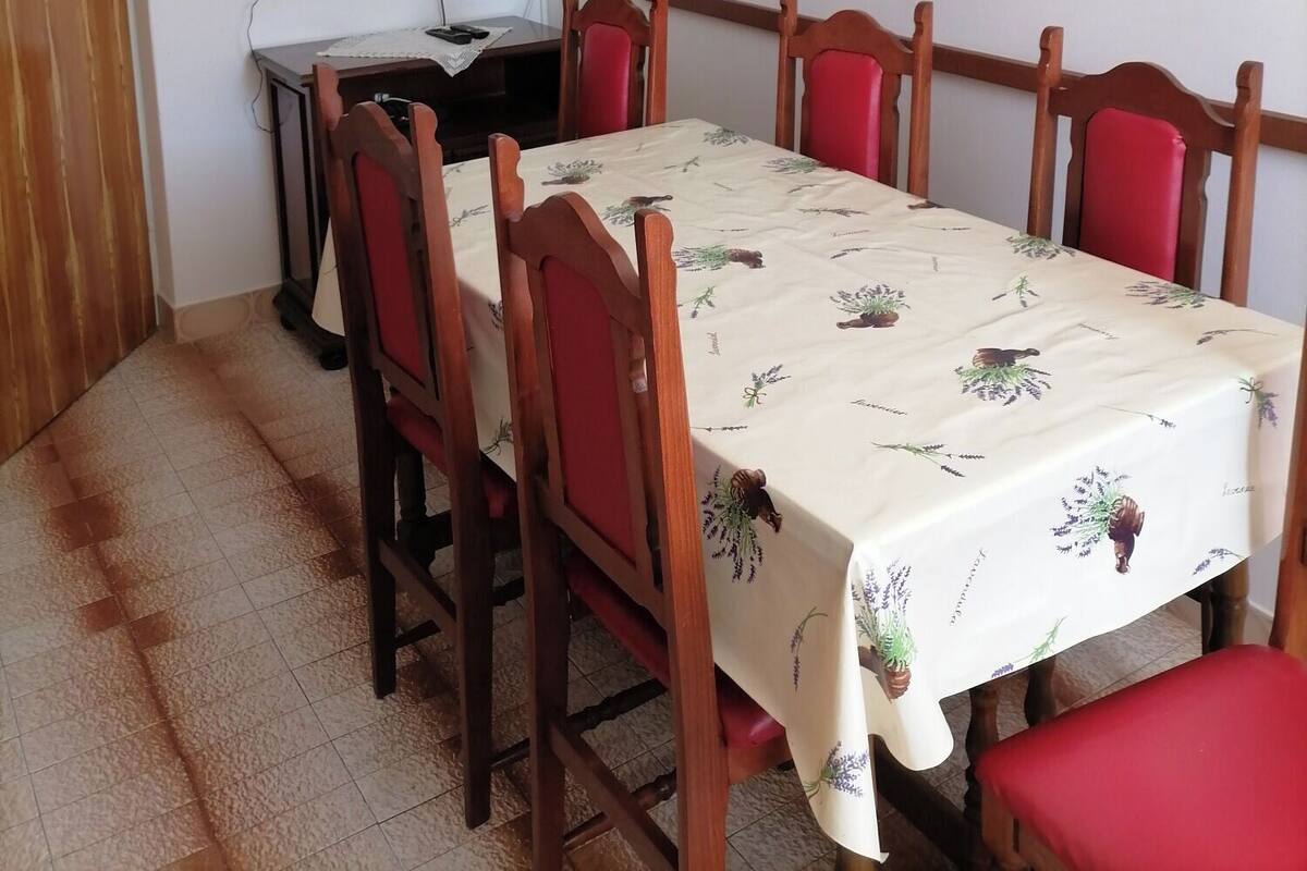 Un comedor con una mesa cubierta por un mantel floral, rodeada de sillas de madera con cojines rojos. Un televisor está montado en la pared sobre la mesa.
