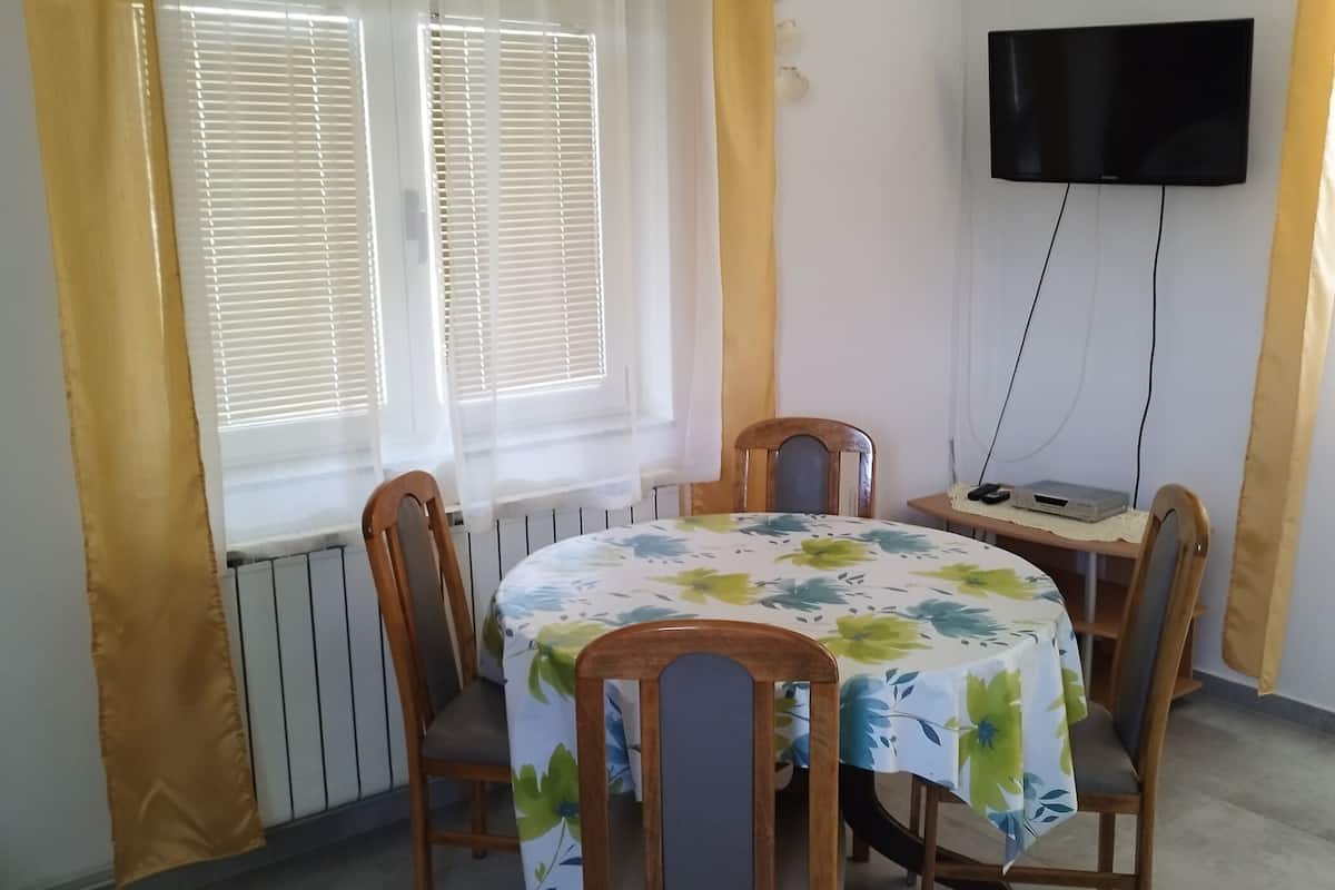 Un comedor con una mesa cubierta con mantel de flores, rodeada de sillas de madera. Hay una ventana con persianas, un equipo de aire acondicionado y un televisor de pantalla plana montado en la pared.