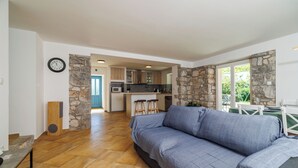 A modern living room with a sofa, a coffee table, and a stone accent wall. A kitchen is visible in the background with wooden cabinets and a microwave. - House Maja (Krk)