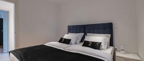 Un dormitorio moderno con una cama grande, ropa de cama blanca y negra, un cabecero oscuro y una mesita de noche con una lámpara.