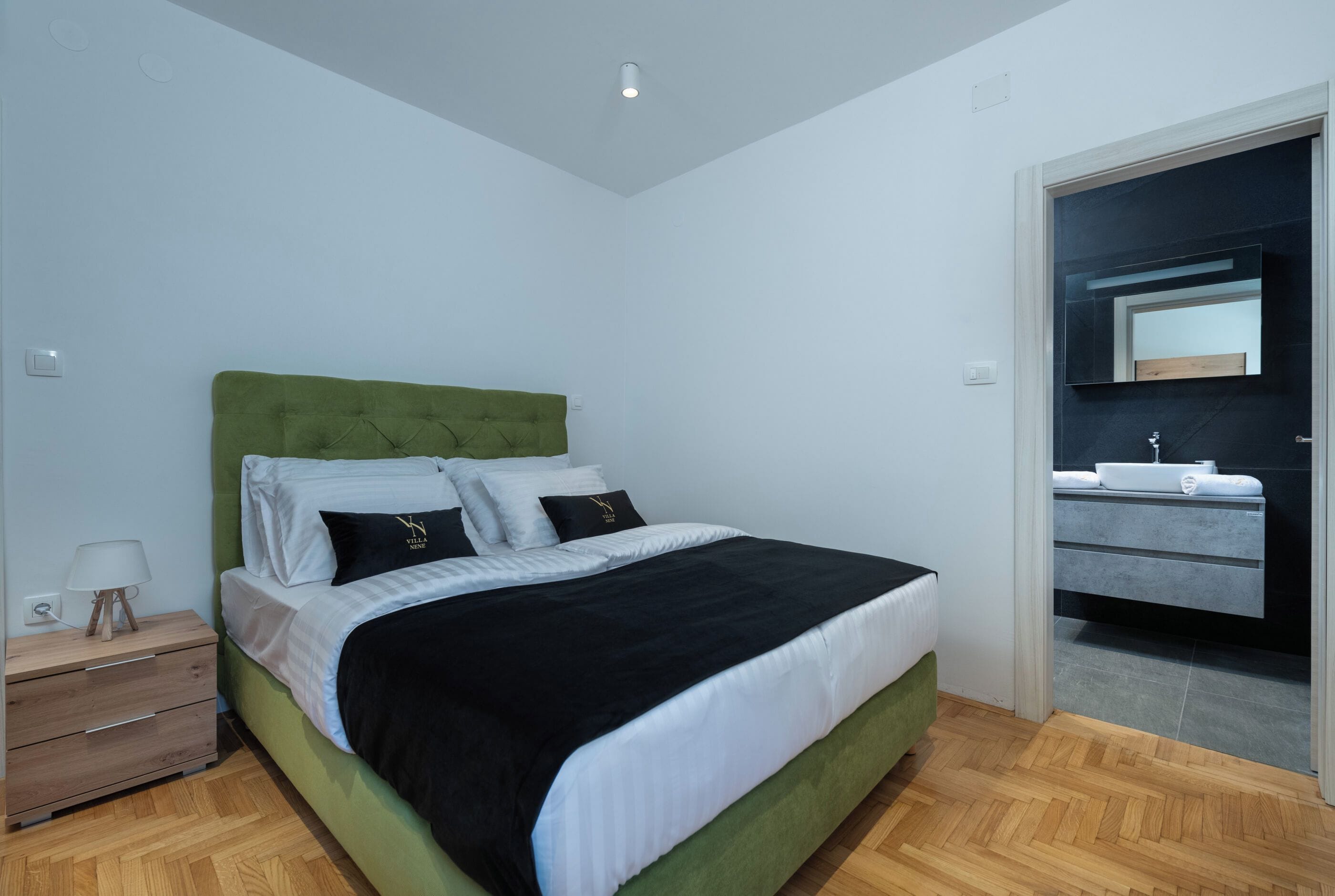 A bedroom with a green upholstered headboard, a wooden nightstand with drawers, a bedside table with a lamp, and a large bed with white and black bedding.