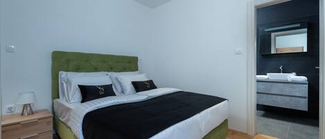 A bedroom with a green upholstered headboard, a wooden nightstand with drawers, a bedside table with a lamp, and a large bed with white and black bedding.