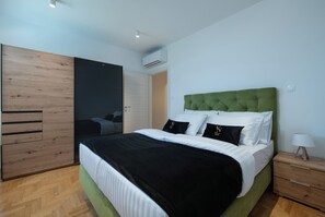 Un dormitorio moderno con un cabecero tapizado en verde, una cama grande con ropa de cama blanca y negra, un armario de madera y una mesita de noche con una lámpara.