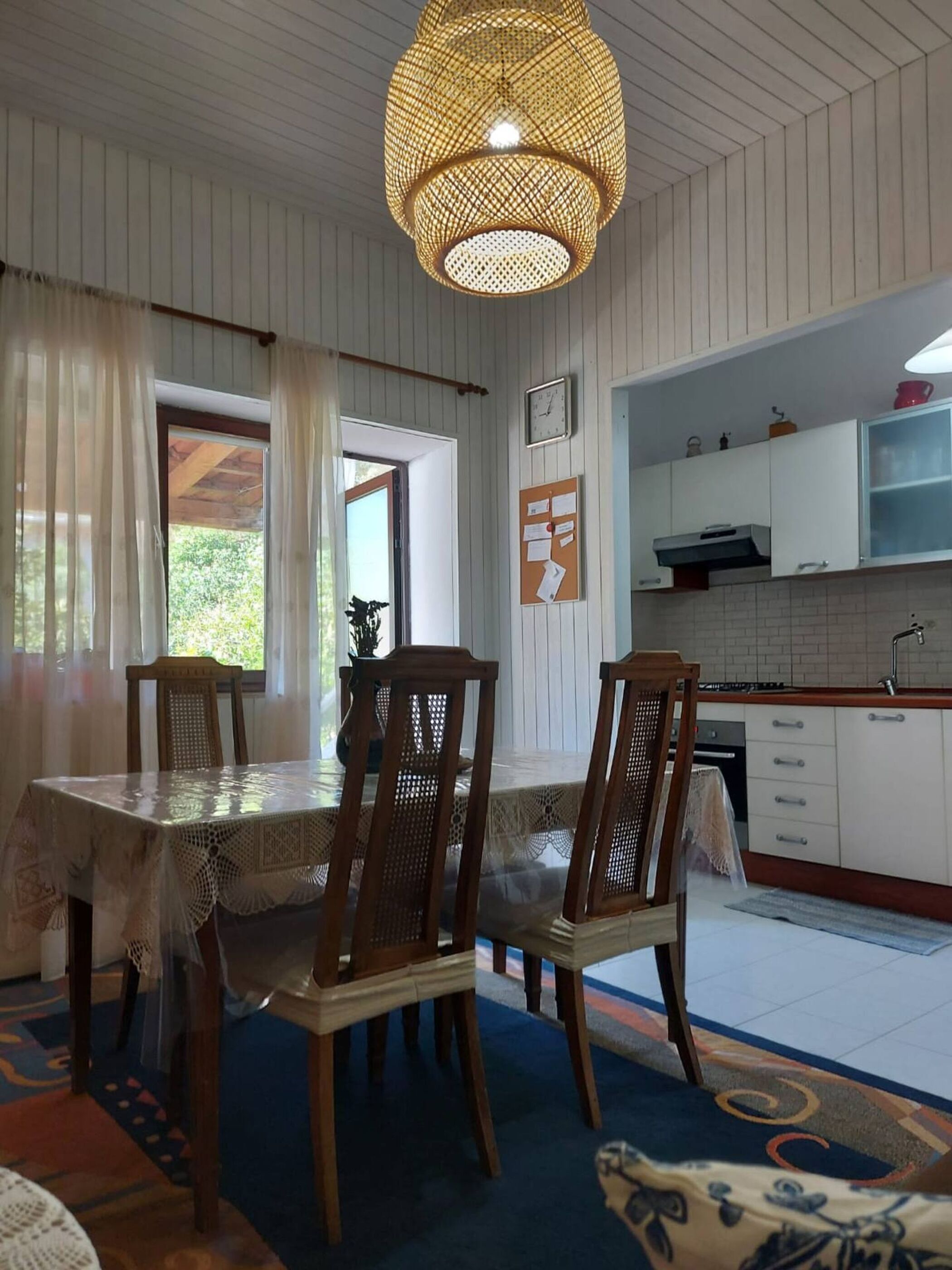 A dining area with a table and chairs, a hanging light fixture, a window with curtains, and a kitchen area with cabinets and a sink.