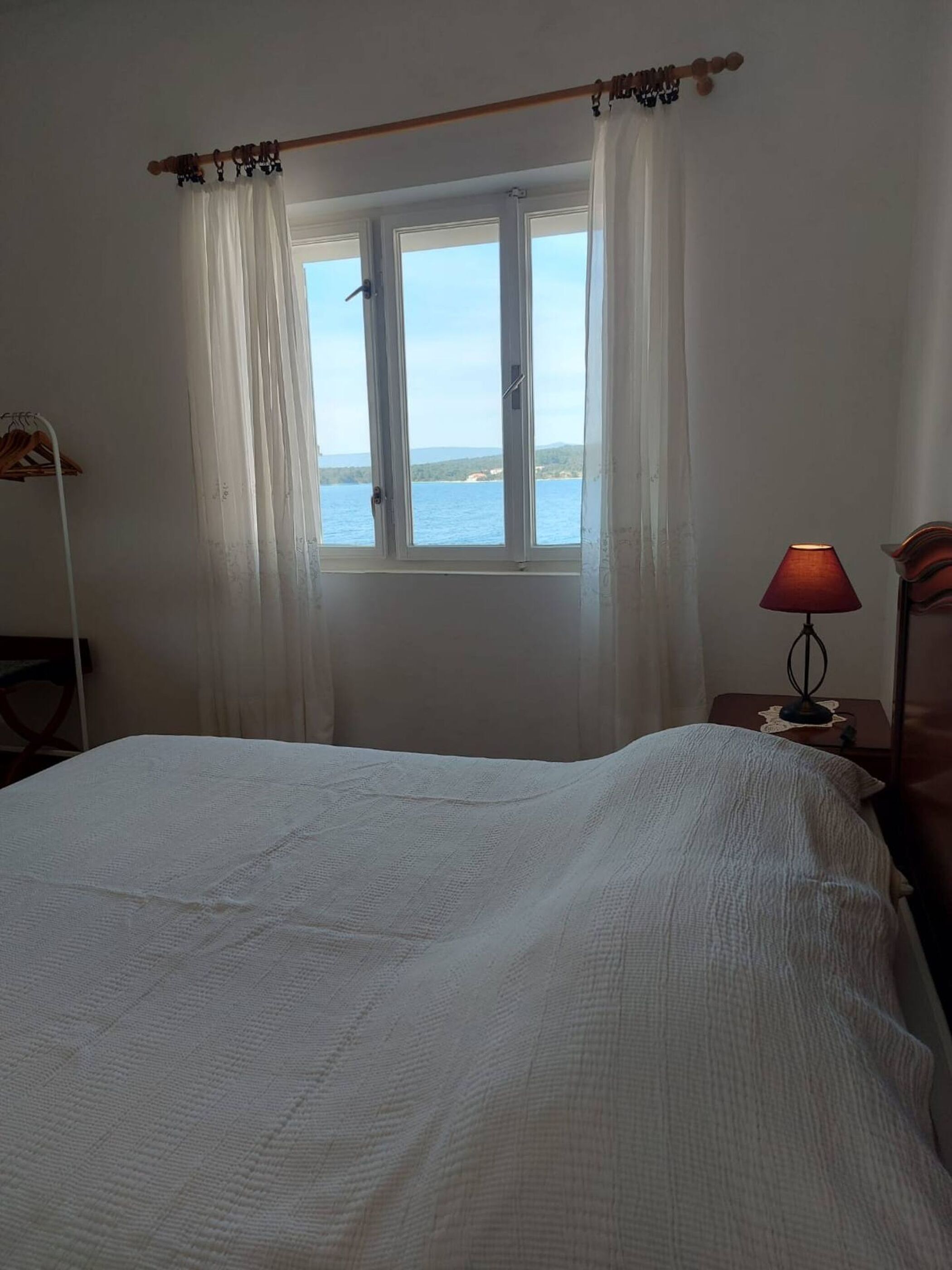 Ein Schlafzimmer mit Bett, einem Fenster mit Meerblick, einer Lampe auf einem Nachttisch und einer Gardinenstange über dem Fenster.
