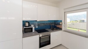 A modern kitchen with white cabinets, a microwave, stove, sink, and a window with a view of a mountain. - Apartment Sankovic (Rovinj)