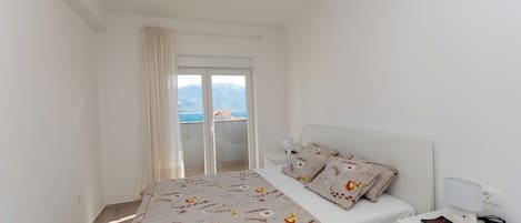 Un dormitorio con una cama, una mesita de noche con una lámpara, un libro y vistas al mar a través de un ventanal corredera.