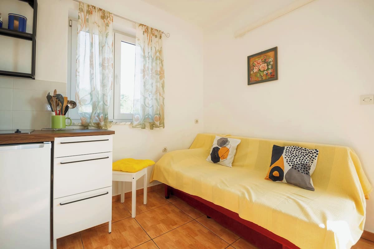 A compact room with a bed, a small table, a chair, and a kitchenette area.