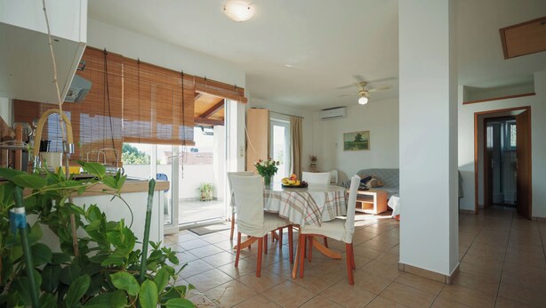 A spacious living area with a dining table, chairs, and a sofa. There is a kitchen area with a stove and cabinets. The room has a large window with blinds and a ceiling fan. - Apartments Kamenar (Dobrinj)