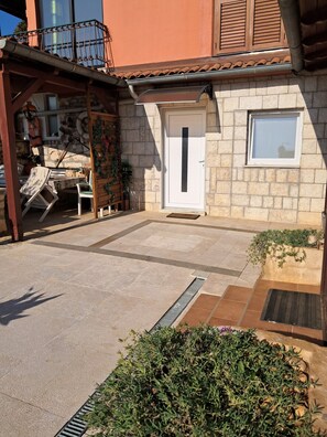 En bygning i sten med et tag af teglsten, en hvid dør og et lille vindue. Der er en overdækket terrasse med en stol og et bord.