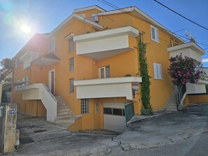 A multi-story building with a yellow facade, a staircase leading to the entrance, and a small garden area with pink flowers. - Apartments Sunflower by the sea (Okrug)